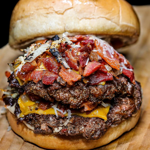 Close-up of a double cheeseburger with bacon, grilled onions, and white sauce on a bun.