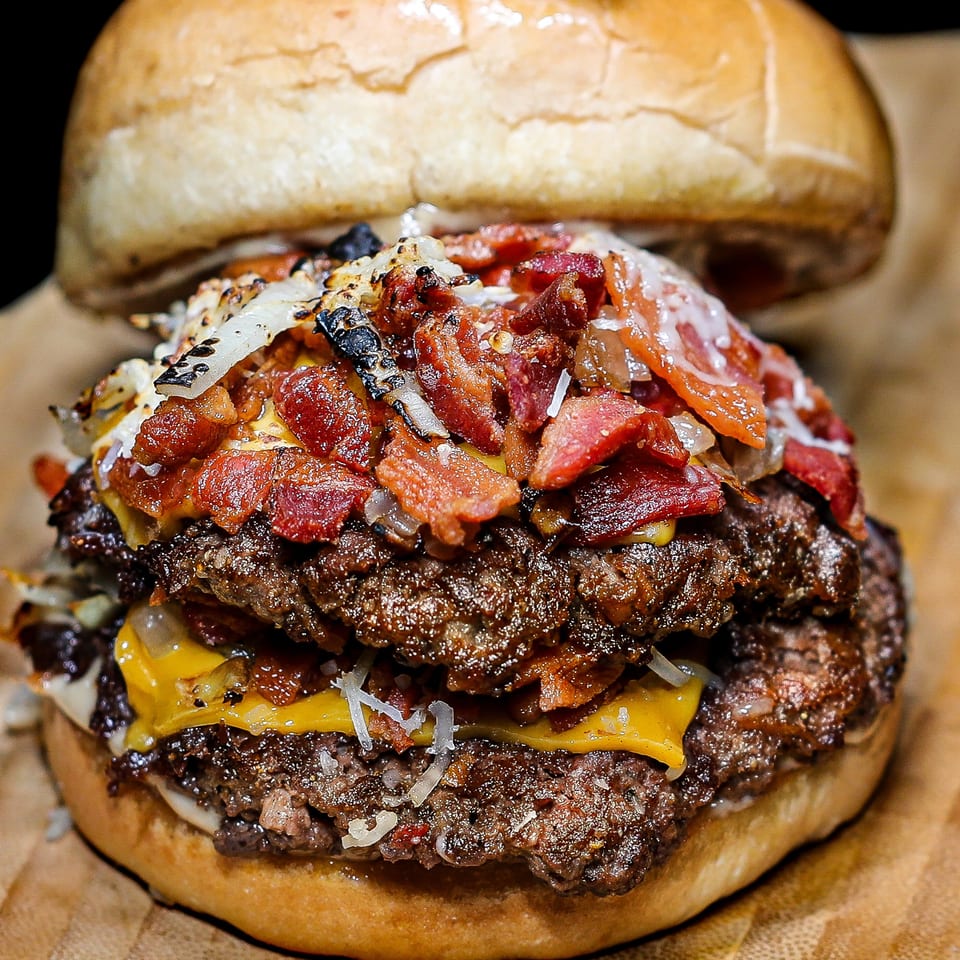Close-up of a double cheeseburger with bacon, grilled onions, and white sauce on a bun.
